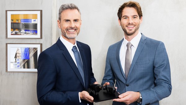 Dvaja muži v oblekoch spoločne držia sochu býka a medveďa – symbolicky predstavujú finančné trhy a partnerskú spoluprácu.