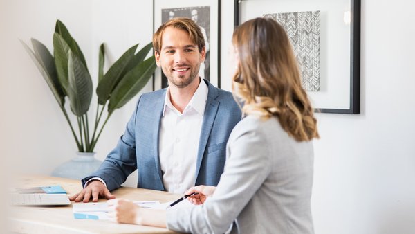 Muž v modrom saku sa priateľsky rozpráva so ženou v svetlom blejzri pri stole.