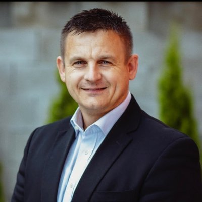 Muž v tmavom obleku a bielej košeli pred stromami