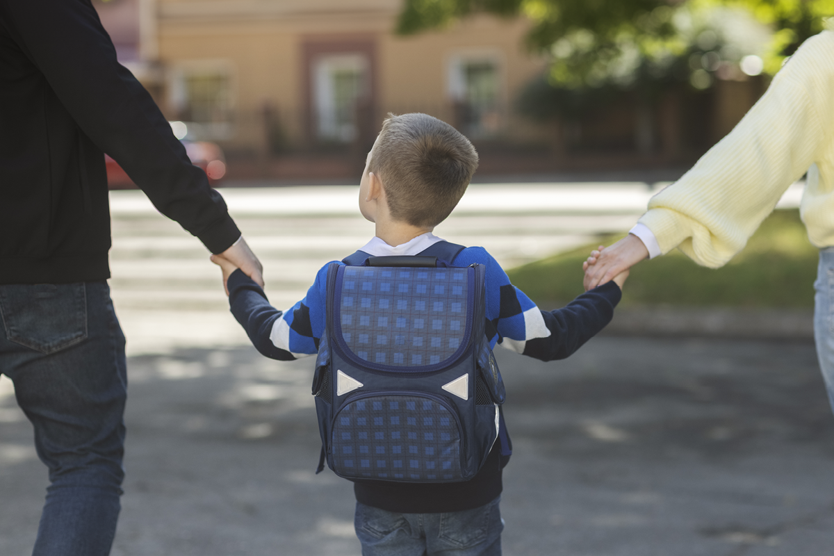 Chlapec s aktovkou drží rodičov za ruky na ceste do školy
