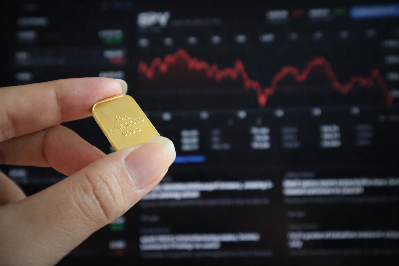 Ruka držiaca malú zlatú tehličku pred monitorom so zobrazeným priebehom finančného trhu.